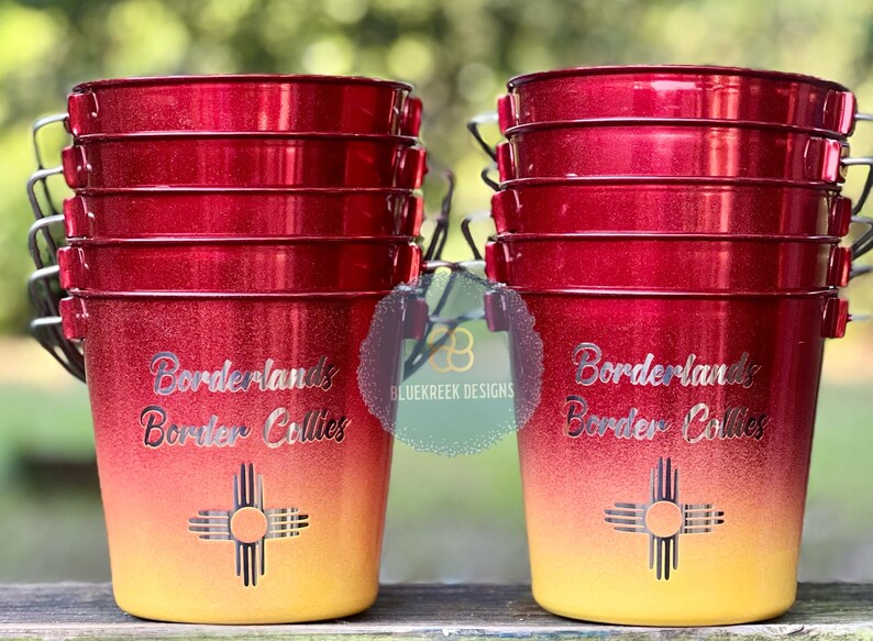May include: Two stacks of red and yellow ombre metal buckets with silver wire handles. Each bucket features the text Borderlands Border Collies and a Zia sun symbol in silver. The finish is glittery, transitioning from deep red at the top to yellow.
