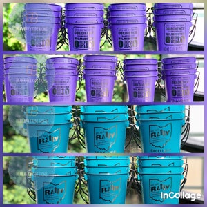 May include: A collection of purple and teal metal buckets featuring printed text and graphics. The purple buckets display 2022 AKC National Obedience Championship Wilmington Ohio, while the teal buckets feature 2022 AKC National Rally Championship Wilmington Ohio.