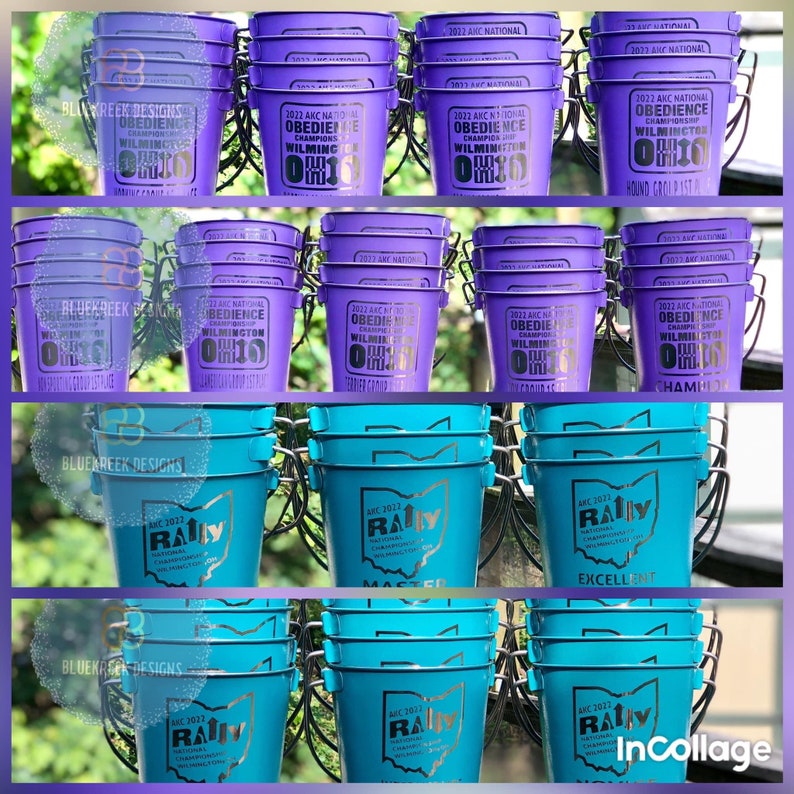 May include: A collection of purple and teal metal buckets featuring printed text and graphics. The purple buckets display 2022 AKC National Obedience Championship Wilmington Ohio, while the teal buckets feature 2022 AKC National Rally Championship Wilmington Ohio.
