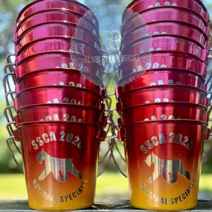 May include: Two stacks of red and yellow gradient metal buckets with silver wire handles. Each bucket features a silver graphic of a dog silhouette, the text SSCA 2026, and the words NATIONAL SPECIALTY printed in a circular arc below the dog.