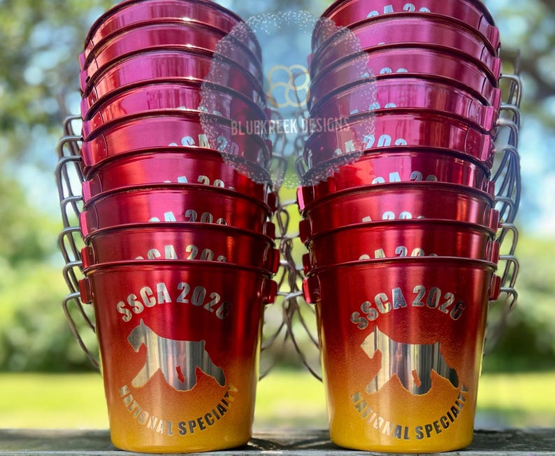 May include: Two stacks of red and yellow gradient metal buckets with silver wire handles. Each bucket features a silver graphic of a dog silhouette, the text SSCA 2026, and the words NATIONAL SPECIALTY printed in a circular arc below the dog.