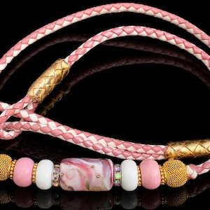 May include: A braided pink and white leather cord necklace featuring a central glass bead with pink and white swirls, flanked by white beads, pink beads, gold-toned textured spherical beads, and two gold-toned cylindrical accents.