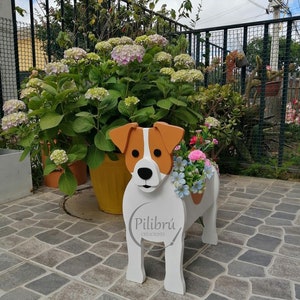 May include: A white wooden dog-shaped planter with brown ears and a brown spot on its back. The planter holds colorful flowers. The text "Pilibrú CREACIONES" is on the side of the planter.