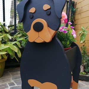 May include: A wooden dog-shaped planter painted black, tan, and brown. The planter is designed to resemble a Rottweiler, with a black body and tan markings. The planter holds pink and purple flowers.
