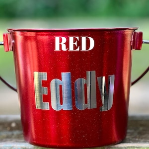 May include: A metallic red bucket with a silver handle and the word RED printed in white at the top. The name Eddy is displayed in large, silver, reflective capital letters across the center of the bucket body, which has a subtle glitter finish.