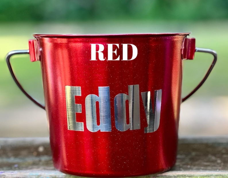 May include: A metallic red bucket with a silver handle and the word RED printed in white at the top. The name Eddy is displayed in large, silver, reflective capital letters across the center of the bucket body, which has a subtle glitter finish.