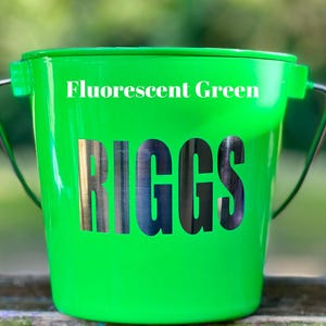 May include: A fluorescent green bucket featuring a metal handle and the name RIGGS in large, bold, dark metallic lettering on the front. The text Fluorescent Green is printed in white above the name on the smooth, glossy surface of the bucket.