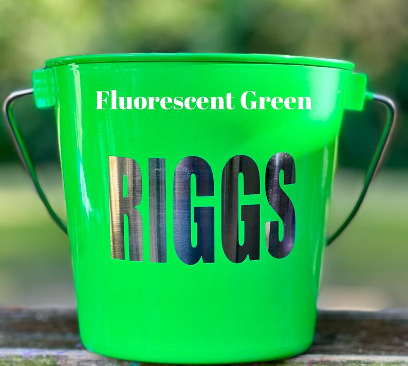 May include: A fluorescent green bucket featuring a metal handle and the name RIGGS in large, bold, dark metallic lettering on the front. The text Fluorescent Green is printed in white above the name on the smooth, glossy surface of the bucket.