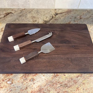May include: A set of three cheese knives with wooden handles and white accents, resting on a dark brown wooden cutting board.