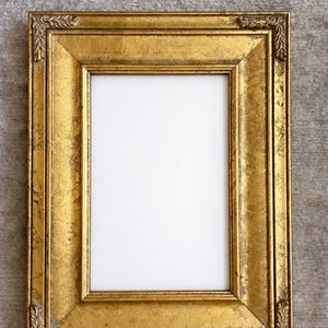 May include: A rectangular picture frame with a distressed gold finish. The frame features a wide, beveled border with decorative leaf-like carvings at each of the four corners. The center of the frame contains a plain, rectangular white insert.