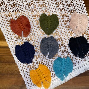 May include: Eight handmade macrame leaf wall hangings in various colors, including orange, green, pink, gray, black, blue, yellow, and navy blue. The hangings are displayed on a white crocheted surface.