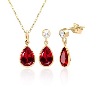 May include: A gold necklace and earrings set with a teardrop-shaped red gemstone pendant and matching earrings. Each earring features a small round clear gemstone above the red gemstone.