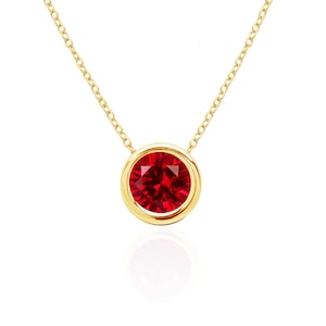 May include: A gold necklace featuring a round, faceted, deep red gemstone set in a smooth, polished gold bezel. The pendant is suspended from a delicate gold chain with fine, interlocking links, creating a simple and elegant jewelry piece.