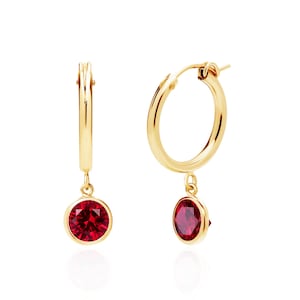 May include: A pair of gold hoop earrings with a dangling, round, deep red gemstone. The earrings are made of polished gold and feature a classic hoop design with a bezel-set ruby-colored stone. The gemstones are approximately 0.3 inches in diameter.