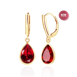 May include: A pair of gold-colored drop earrings with teardrop-shaped, deep red gemstones. The earrings have a lever-back closure and are displayed against a white background. A "NEW COLLECTION" sticker is visible.