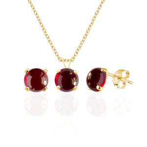 May include: A jewelry set consisting of a gold chain necklace with a round, deep red gemstone pendant and a pair of matching round, deep red gemstone stud earrings. Each piece features a polished gold setting with four prongs holding the stones.