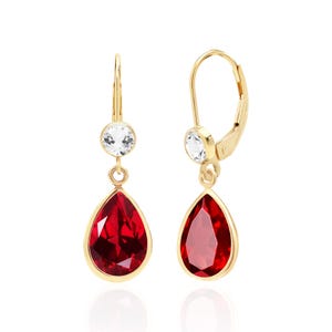 May include: A pair of gold-colored drop earrings. Each earring features a teardrop-shaped, faceted red gemstone suspended below a round, clear gemstone. The earrings have a lever-back closure.
