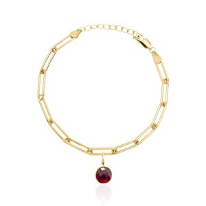 May include: A gold-colored link bracelet with a lobster clasp closure. The bracelet features a small, round, faceted, red gemstone pendant. The jewelry is displayed against a white background.