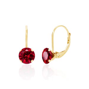 May include: A pair of gold leverback earrings, each featuring a single round-cut, deep red gemstone held in a four-prong setting. The metal has a polished yellow gold finish, and the design includes a secure hinged closure mechanism.
