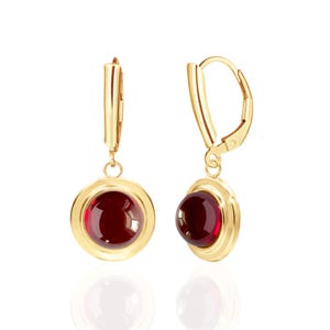 May include: A pair of gold-colored leverback earrings. Each earring features a round, deep red gemstone set within a gold bezel. The earrings are displayed against a white background, with a slight reflection visible below.