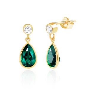 May include: A pair of gold-colored drop earrings. Each earring features a teardrop-shaped, emerald-green gemstone framed in gold, suspended below a small, round, clear gemstone. The earrings are set against a white background.