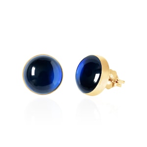 May include: A pair of stud earrings featuring round, deep blue cabochon stones set in polished gold-tone metal. One earring is shown from the front, while the other is angled to display the side profile and the post backing mechanism.