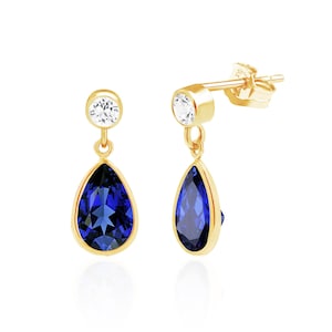 May include: A pair of gold drop earrings featuring a round clear stone at the top and a larger pear-shaped deep blue stone suspended below. The stones are set in gold bezels, and the earrings include a post-back closure for secure attachment.
