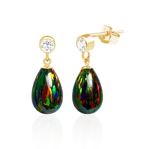 May include: A pair of gold-colored drop earrings. Each earring features a clear, round stone at the top, connected to a teardrop-shaped, iridescent black and green stone. The earrings are set against a white background.