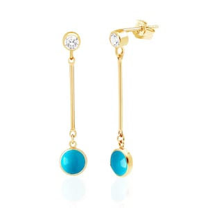 May include: A pair of gold drop earrings. Each earring has a small, clear gemstone at the top, a long, thin gold bar, and a round, turquoise stone at the bottom. The earrings are set against a white backdrop.