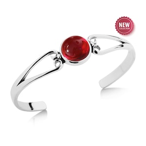 May include: A silver cuff bracelet with a large, round, red gemstone at the center. The bracelet has an open design with decorative flourishes near the gemstone. A "NEW COLLECTION" sticker is in the upper right corner.