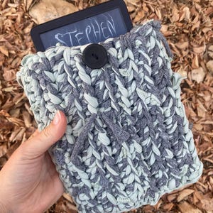 以下が含まれることがあります： グレーとライトグリーンのかぎ針編みタブレットスリーブ、黒いボタン留め。タブレット画面には白で「STEPHEN」という名前が表示されています。スリーブは太く、質感のある糸で作られています。