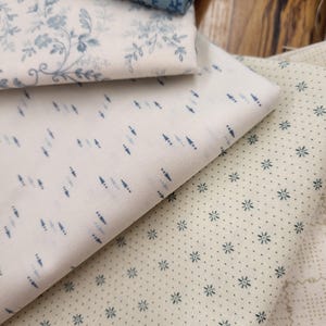 May include: A collection of folded fabric pieces featuring various patterns. One piece has a blue and white floral print, another displays a white background with small blue dash marks, and a third shows a cream background with tiny blue stars.