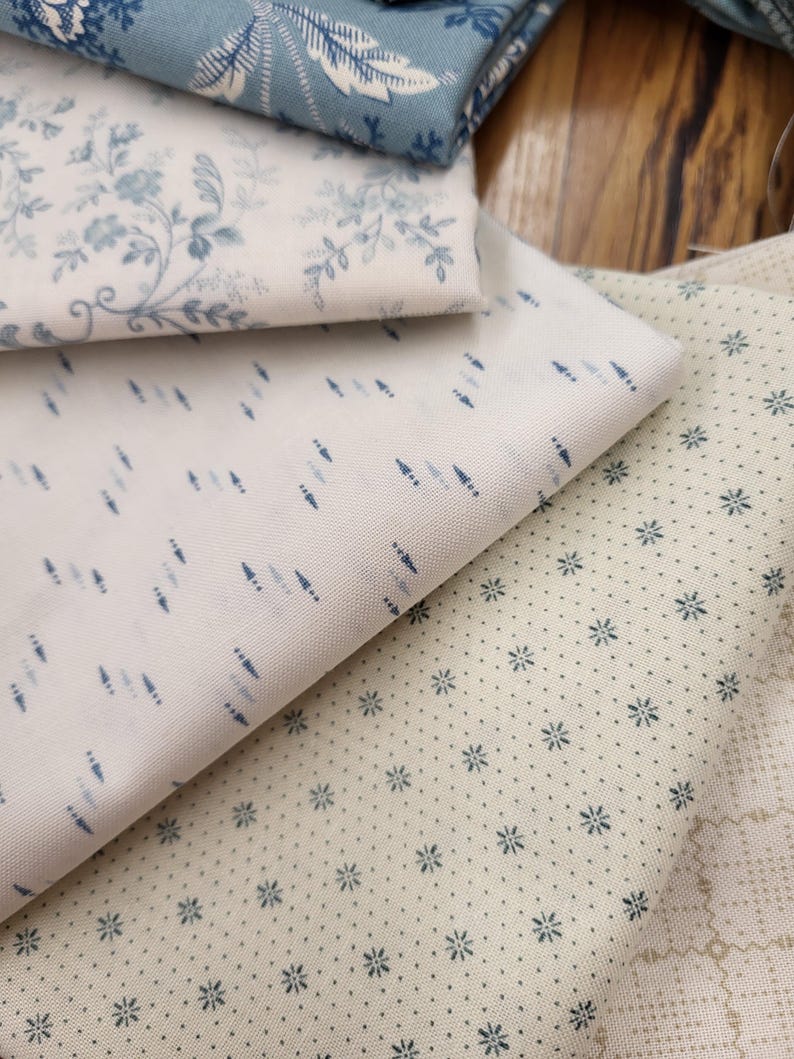 May include: A collection of folded fabric pieces featuring various patterns. One piece has a blue and white floral print, another displays a white background with small blue dash marks, and a third shows a cream background with tiny blue stars.