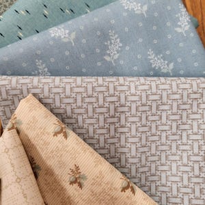 May include: A collection of folded fabric swatches featuring various patterns. The stack includes a light blue fabric with white floral prints, a blue fabric with dark dashes, a beige fabric with a woven grid, and a beige fabric with small floral motifs.