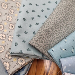 May include: A collection of folded fabric swatches in shades of light blue, grey, and white. The patterns include small floral prints, delicate leaf motifs, and geometric designs. Each piece of fabric displays a distinct texture and color palette.