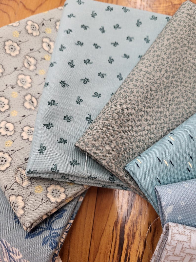 May include: A collection of folded fabric swatches in shades of light blue, grey, and white. The patterns include small floral prints, delicate leaf motifs, and geometric designs. Each piece of fabric displays a distinct texture and color palette.