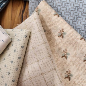 May include: A collection of folded fabric swatches featuring various patterns. The textiles include cream and beige backgrounds with small blue floral prints, a tan grid pattern, and a light blue fabric with a woven texture and floral motifs.