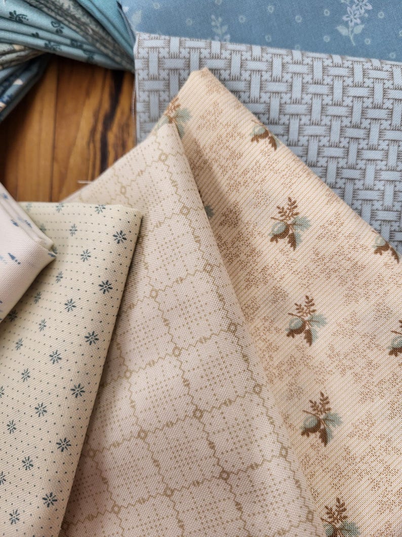 May include: A collection of folded fabric swatches featuring various patterns. The textiles include cream and beige backgrounds with small blue floral prints, a tan grid pattern, and a light blue fabric with a woven texture and floral motifs.