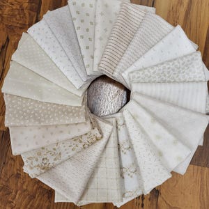 May include: A collection of folded fabric squares arranged in a circular fan pattern. Each piece of fabric features a unique off-white, cream, or beige color palette with various printed patterns including floral, geometric, striped, and dotted designs.