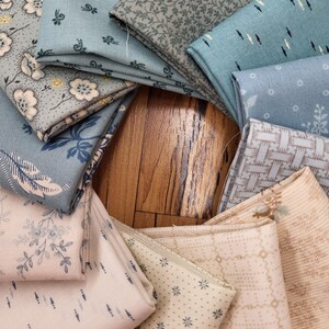 May include: A collection of folded fabric squares arranged in a circular pattern. The textiles feature various patterns including floral prints, geometric shapes, small dots, and woven textures in a color palette of soft blues, creams, and tans.