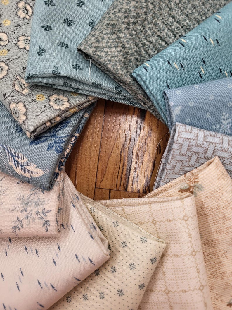 May include: A collection of folded fabric squares arranged in a circular pattern. The textiles feature various patterns including floral prints, geometric shapes, small dots, and woven textures in a color palette of soft blues, creams, and tans.