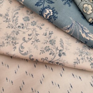 May include: Three folded pieces of fabric featuring distinct patterns. One piece is cream with blue floral vines, another is a solid dusty blue with white floral motifs, and the third is a cream fabric with small, vertical blue dash patterns.