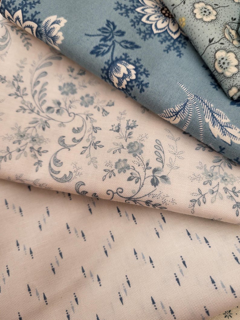 May include: Three folded pieces of fabric featuring distinct patterns. One piece is cream with blue floral vines, another is a solid dusty blue with white floral motifs, and the third is a cream fabric with small, vertical blue dash patterns.