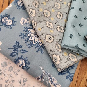 May include: Four folded pieces of fabric in shades of blue and white. One piece features a dark blue floral pattern on a light blue background, another has white flowers with yellow centers, one shows a small dark blue sprig print, and one is white.
