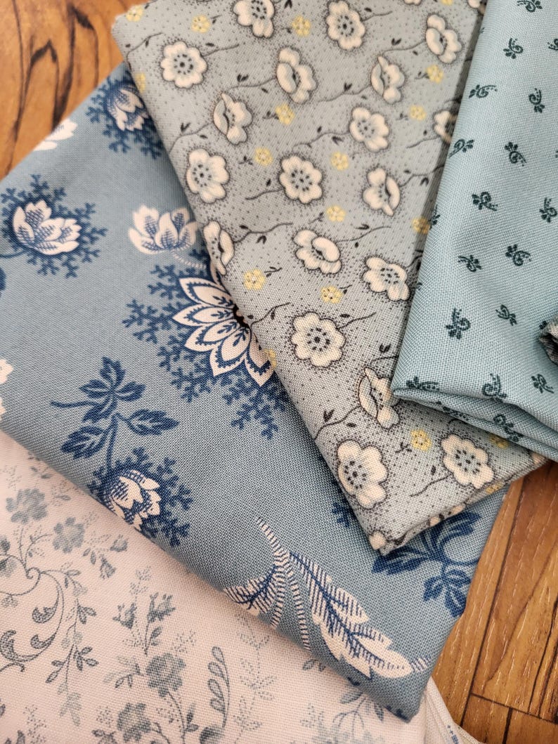 May include: Four folded pieces of fabric in shades of blue and white. One piece features a dark blue floral pattern on a light blue background, another has white flowers with yellow centers, one shows a small dark blue sprig print, and one is white.