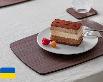 May include: A wooden placemat with a chocolate cake with white and brown layers on a white plate with raspberries and yellow fruit. The placemat is on a white tablecloth.
