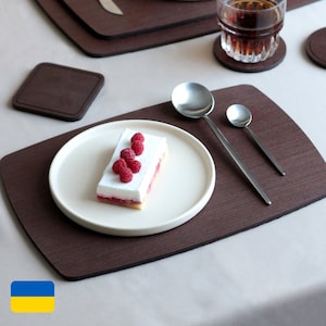 Könnte beinhalten: Ein weißer Teller mit einem Stück Kuchen, das mit Himbeeren belegt ist, steht auf einem braunen Holz-Untersetzer. Zwei silberne Löffel liegen rechts neben dem Teller auf dem Untersetzer.