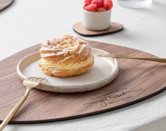 May include: A wooden serving tray with a rounded triangular shape. The tray has a white ceramic plate with a pastry on it. The tray has the words "The best moments are those gathered around the table" engraved on it.