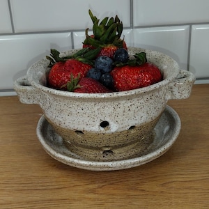 Könnte beinhalten: Ein weißer Keramik-Abtropfkorb mit braunen Flecken und zwei Griffen, gefüllt mit frischen Erdbeeren und Blaubeeren. Der Abtropfkorb steht auf einem passenden weißen Keramikteller mit braunen Flecken.