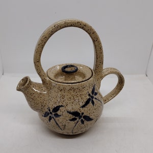 Puede incluir: Una tetera de cerámica beige con un esmalte moteado y diseños florales azules. La tetera tiene un asa única que se curva sobre la parte superior.